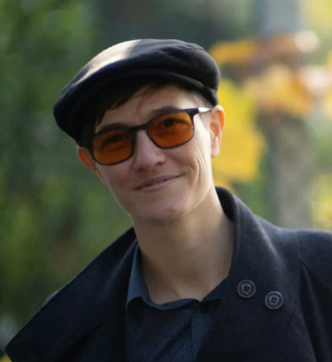 fuzzy headshot showing a white person smiling at the camera, wearing a black hat, yellow-tinted glasses, blue collard shirt, and grey coat