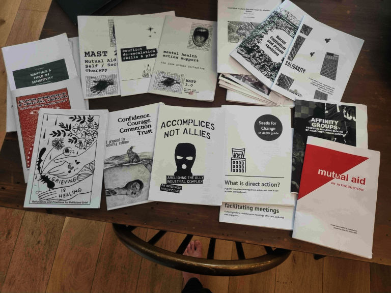 a photo of a collection of zines spread haphazardly on a wooden table, a bare white foot is visible through the rungs of the back of a chair